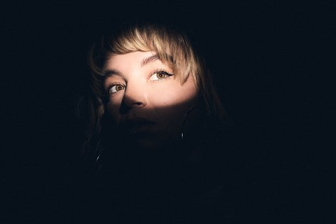A close-up portrait of a person with light illuminating part of their face, creating a contrast against a dark background. Their gaze is directed slightly upward, conveying a sense of intrigue or contemplation.
