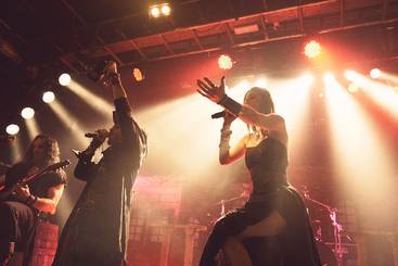 Eine Sängerin und ein Sänger stehen auf der Bühne und performen vor einem Publikum. Im Hintergrund sind Musiker mit Instrumenten zu sehen. Die Szene wird von Bühnenlichtern beleuchtet, die eine energische Atmosphäre schaffen.