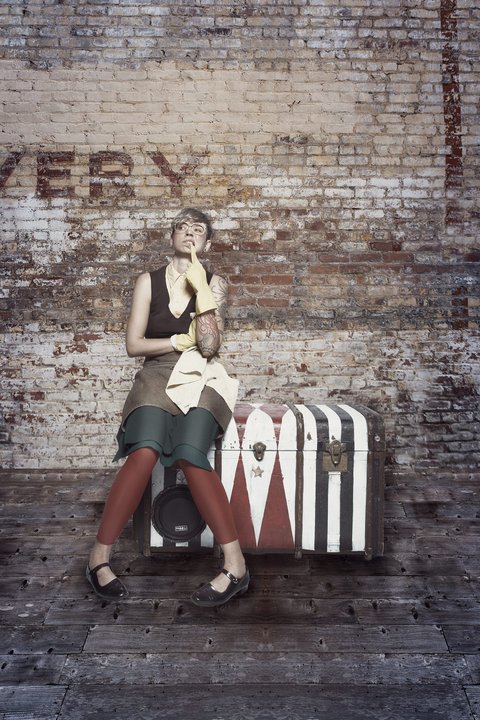 Eine lächelnde Frau sitzt auf einer auffälligen Truhe mit rot-weißen Streifen. Sie trägt ein modisches Outfit in Erdtönen und hat einen gelben Schal. Der Hintergrund ist eine rustikale Ziegelwand. Die Szene vermittelt eine entspannte und freundliche Atmosphäre.