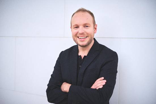 Ein lächelnder Mann steht mit verschränkten Armen vor einer hellen Wand. Er trägt einen dunklen Anzug und einen schwarzen Pullover darunter. Der Ausdruck auf seinem Gesicht vermittelt Freundlichkeit und Selbstbewusstsein.