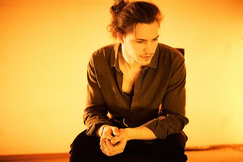 A young man sits pensively, wearing a dark shirt. He has long hair tied back and a thoughtful expression, with warm orange lighting creating a soft atmosphere. His hands are clasped on his knees, and he appears deep in contemplation.