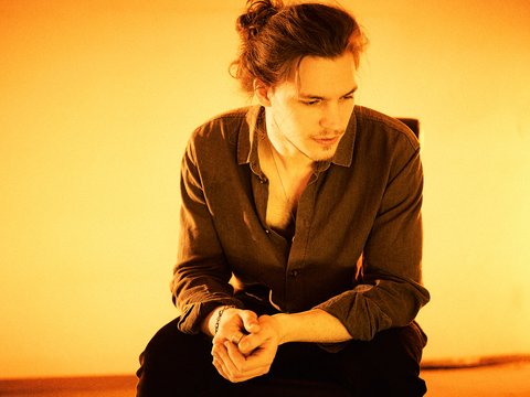 Ein junger Mann sitzt nachdenklich auf einem Stuhl. Er trägt ein dunkles Hemd und hat längere, zum Zopf gebundene Haare. Der Hintergrund ist in warmen Orangetönen gehalten, was eine ruhige und intime Stimmung vermittelt.