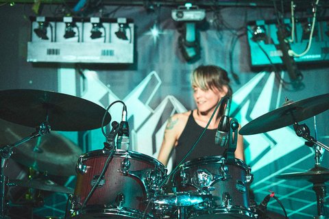 Eine Frau sitzt hinter einem Schlagzeug und spielt. Sie hat kurze Haare und trägt ein schwarzes Oberteil. Im Hintergrund sind Lichtelemente und eine grafische Wanddekoration, die zur Rock-Atmosphäre des Auftritts beitragen.