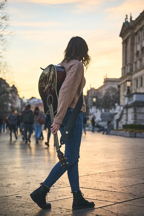 Eine Person mit einer Gitarre auf dem Rücken geht auf einem Stadtweg, während die Sonne untergeht. Die Kleidung ist lässig und die Umgebung zeigt andere Menschen im Hintergrund. Die Szene vermittelt ein Gefühl von Entspannung und musikalischer Gemeinschaft.