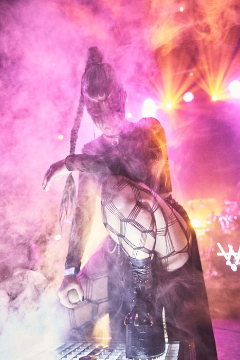 A performer with an elaborate costume stands amidst colorful smoke and dramatic lighting during a live show. The outfit features intricate designs, emphasizing creativity and theatricality. The atmosphere is lively and energetic, capturing the essence of a captivating performance.