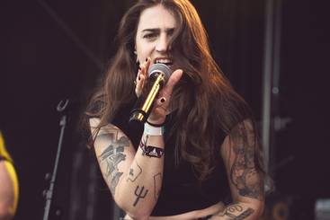 Vulvarine Eine Frau mit langen, offenen Haaren singt leidenschaftlich ins Mikrofon. Sie trägt ein schwarzes Top und hat viele Tattoos auf ihren Armen. Die Szene vermittelt Energie und Intensität einer Live-Musikaufführung.