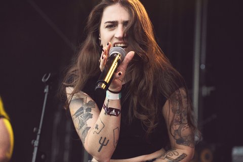 A female singer with long hair passionately performs on stage, holding a microphone. She has visible tattoos on her arms and appears engaged with the audience, expressing strong emotion through her performance. The background hints at a musical event atmosphere.