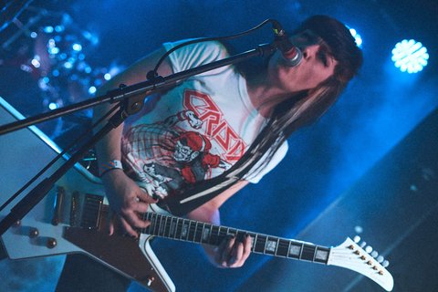 Eine Musikerin mit langen, dunklen Haaren spielt Gitarre auf einer Bühne. Sie trägt ein T-Shirt mit einem grafischen Druck und singt ins Mikrofon, während bunte Lichter im Hintergrund leuchten.