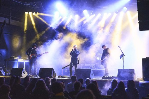 Ein Live-Konzert mit einer Band auf der Bühne. Die Musiker spielen Instrumente, während bunte Lichter und Nebel die Atmosphäre prägen. Im Vordergrund sind zahlreiche Zuschauer, die der Musik aufmerksam folgen.