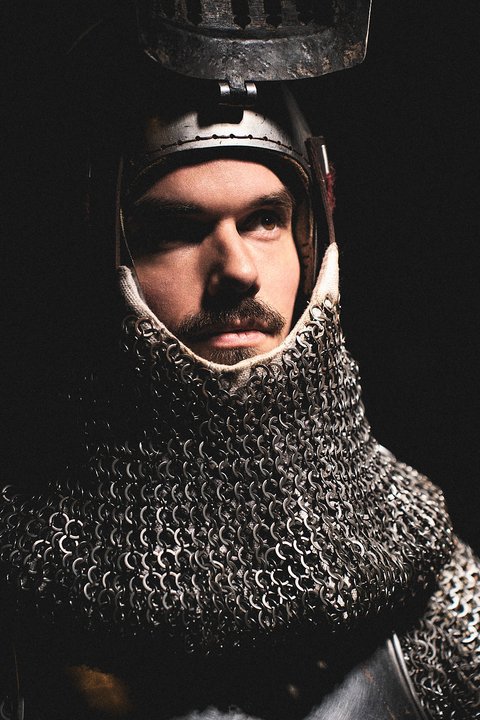 Ein Mann trägt eine mittelalterliche Rüstung aus Kettenhemd und Helm. Sein ernster Gesichtsausdruck und die dunkle Umgebung verleihen ihm eine kämpferische Ausstrahlung. Die Bildkomposition konzentriert sich auf den Kopf und Oberkörper des Ritters.