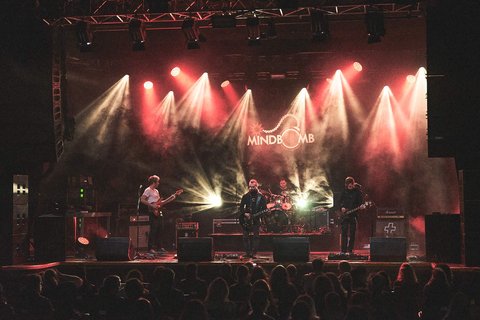 Eine Band tritt auf einer Bühne auf, umgeben von bunten Lichtern und Nebel. Die Musiker spielen Instrumente und stehen im Vordergrund, während das Publikum im Hintergrund zuschaut. Das Bandlogo "MIND BOMB" ist sichtbar hinter ihnen.