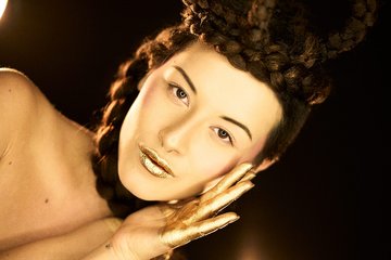 Eine Frau mit kunstvollem Zopf trägt goldenen Lippenstift und hat goldene Akzente auf ihren Händen. Ihr Gesicht drückt eine sanfte Eleganz aus, während sie mit einer Hand ihr Gesicht berührt. Der Hintergrund ist dunkel, was die Gesichtszüge betont.