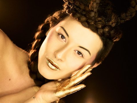 Eine Frau mit kunstvollem Zopf trägt goldenen Lippenstift und hat goldene Akzente auf ihren Händen. Ihr Gesicht drückt eine sanfte Eleganz aus, während sie mit einer Hand ihr Gesicht berührt. Der Hintergrund ist dunkel, was die Gesichtszüge betont.