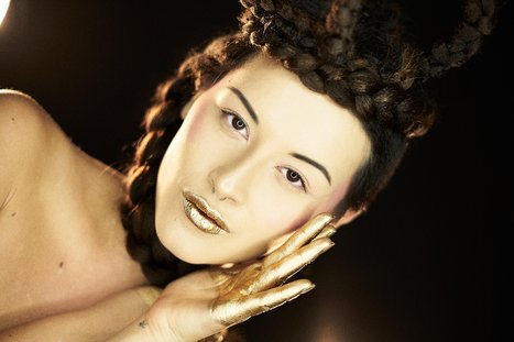 A close-up portrait of a person with intricate braided hair and golden makeup accents. They are posing gracefully, with one hand touching their face, conveying a sense of elegance and artistic expression against a dark background.