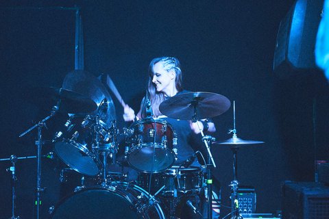 Eine Drummerin spielt energetisch an einem Schlagzeug auf der Bühne. Ihr Gesicht strahlt Freude aus, während sie im Scheinwerferlicht steht. Der Hintergrund ist dunkel, was den Fokus auf die Musikerin und ihr Instrument legt.