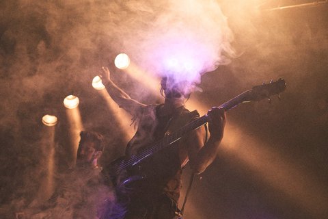 Ein Musiker spielt E-Bass auf einer Bühne, umgeben von dichter Nebelkulisse und warmen Lichtstrahlen. Eine zweite Person ist im Hintergrund zu sehen, während die Atmosphäre eine energetische Live-Performance vermittelt.