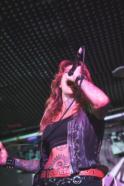 Eine weibliche Sängerin mit tätowierten Armen steht auf der Bühne und hält ein Mikrofon. Sie trägt eine schwarze Lederjacke und einen roten Gürtel. Die Atmosphäre ist energiegeladen und es ist Dunkelheit im Hintergrund zu sehen.