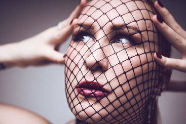 Eine Frau mit einem Netzschleier, der ihr Gesicht teilweise bedeckt. Sie hat einen konzentrierten Ausdruck und hält sich mit beiden Händen an Kopf und Gesicht. Ihre Lippen sind rot lackiert, und ihr Blick ist nachdenklich. Der Hintergrund ist neutral.