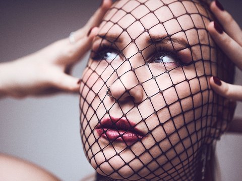 Eine Frau mit einem Netzschleier, der ihr Gesicht teilweise bedeckt. Sie hat einen konzentrierten Ausdruck und hält sich mit beiden Händen an Kopf und Gesicht. Ihre Lippen sind rot lackiert, und ihr Blick ist nachdenklich. Der Hintergrund ist neutral.