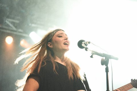 Eine Sängerin steht auf der Bühne und performt. Sie trägt ein schwarzes Oberteil und hat lange, offene Haare. Ihr Gesicht ist fröhlich, während sie ins Mikrofon singt. Im Hintergrund sind helles Licht und unscharfe Bühnenelemente zu erkennen.