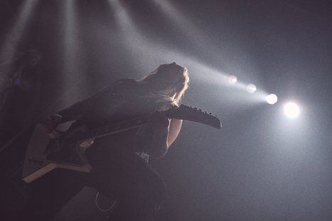 A silhouetted musician plays an electric guitar on stage, illuminated by dramatic lighting from above. The scene conveys energy and intensity, capturing the essence of a live performance.