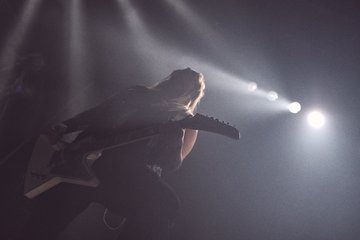 Ein Musiker spielt leidenschaftlich Gitarre auf der Bühne. Starke Lichteffekte und Nebel schaffen eine dynamische Atmosphäre. Die Silhouette des Musikers hebt sich dramatisch vor den Lichtern ab und vermittelt ein Gefühl von Energie und Intensität.