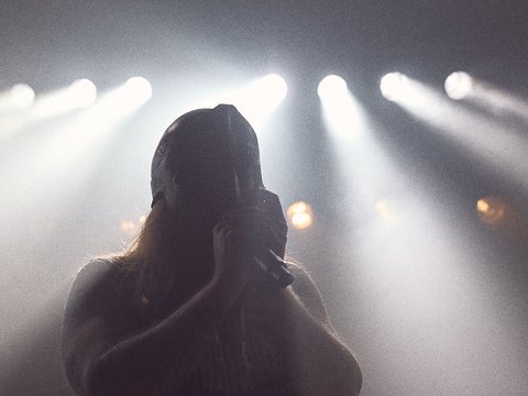 Eine silhouettenhafte Person hält ein Mikrofon, umgeben von schimmerndem Licht von einem Bühnenlicht. Die Atmosphäre wirkt intensiv und energiegeladen, was auf eine musikalische Darbietung hindeutet.