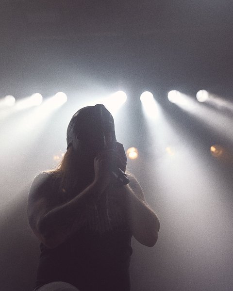 A silhouette of a performer holding a microphone, illuminated by bright stage lights. The focus is on the figure, creating a dramatic atmosphere, with beams of light radiating from above. The scene conveys an energetic live music performance.
