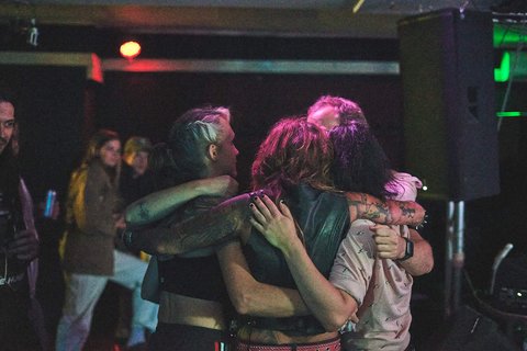 Vier Menschen umarmen sich in einer festlichen Umgebung. Sie stehen eng zusammen, lächelnd und in einer herzlichen Atmosphäre. Im Hintergrund sind weitere Personen zu sehen, die an einer Veranstaltung teilnehmen. Die Szene strahlt Freude und Gemeinschaft aus.