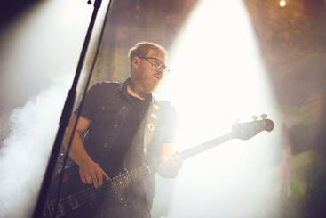 Ein Musiker spielt Bass auf einer Bühne. Er trägt eine dunkle Weste und Brille. Im Hintergrund ist helles Licht und Nebel zu sehen, was eine stimmungsvolle Atmosphäre schafft.