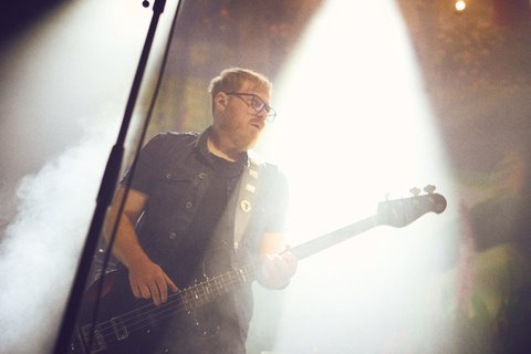Ein Musiker spielt Bass auf einer Bühne. Er trägt eine dunkle Weste und Brille. Im Hintergrund ist helles Licht und Nebel zu sehen, was eine stimmungsvolle Atmosphäre schafft.