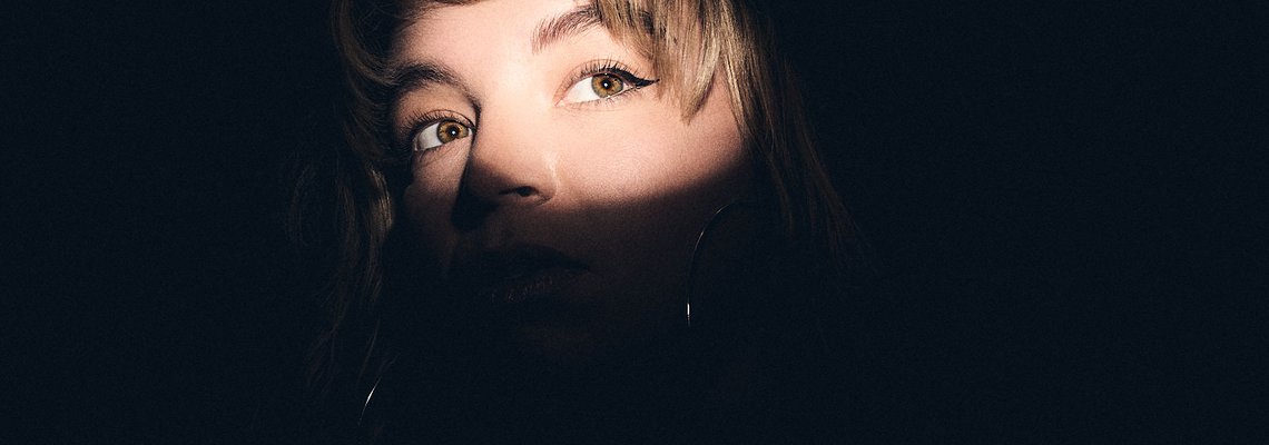 Eine Person mit kurzen, leicht gewellten Haaren schaut mit einem nachdenklichen Ausdruck. Ihr Gesicht wird teilweise von Schatten bedeckt, während ein Lichtstrahl ihr Auge betont. Der Hintergrund ist dunkel, was die Aufmerksamkeit auf ihren Blick lenkt.