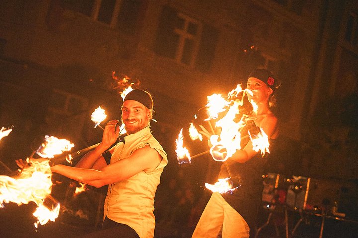 Zwei Performer führen eine beeindruckende Feuershow auf. Der männliche Artist hält brennende Fackeln und lächelt, während die weibliche Künstlerin ebenfalls brennende Objekte präsentiert. Im Hintergrund ist eine dunkle Umgebung mit verschwommenen Lichtern zu sehen.