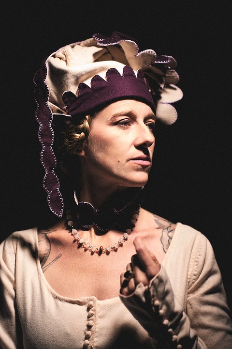 Eine Frau in historischer Kleidung mit einer eleganten Kopfbedeckung und einer Perlenkette blickt nachdenklich zur Seite. Der Hintergrund ist dunkel, wodurch ihr Gesicht und die Details ihrer Kleidung hervorgehoben werden.