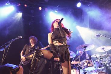 Eine Sängerin mit rotem Haar steht auf der Bühne und hält ein Mikrofon. Sie trägt ein schwarzes, glitzerndes Outfit und hat eine dynamische Pose. Im Hintergrund sind zwei Musiker am Schlagzeug und an der Gitarre zu sehen, beleuchtet von bunten Lichtern.