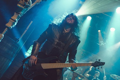 Ein Musiker mit langen, lockigen Haaren und einem schwarzen Outfit spielt Bass auf einer Bühne. Im Hintergrund sind weitere Musiker zu sehen, während Lichter und ein mystisches Ambiente die Szene beleuchten. Der Musiker hat einen intensiven Ausdruck und zeigt seine Leidenschaft für die Musik.