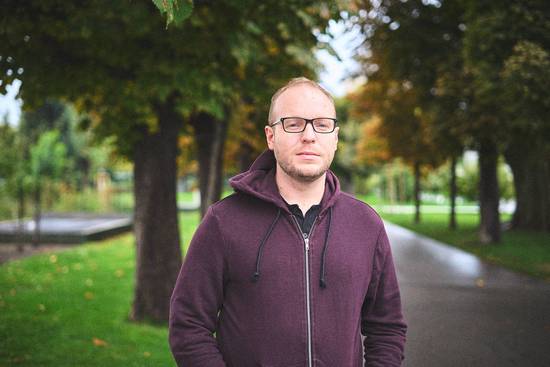Ein Mann mit Brille steht in einem Park. Er trägt einen lila Kapuzenpullover und blickt ernst in die Kamera. Im Hintergrund sind grüne Bäume und ein Weg zu sehen, der schmal und gepflastert ist.