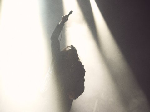Eine silhouettierte Person steht auf einer Bühne und hebt ein Mikrofon, umgeben von dramatischem Licht und Nebel. Die Szene vermittelt eine intensive, musikalische Atmosphäre.