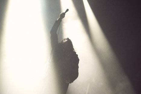 A silhouetted performer stands with one arm raised, holding a microphone, illuminated by bright stage lights. The background is hazy, enhancing the dramatic effect of the moment.