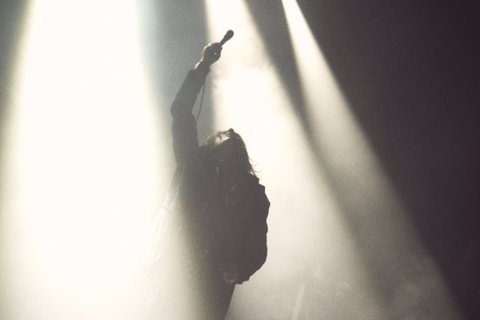 A silhouetted performer stands with one arm raised, holding a microphone, illuminated by bright stage lights. The background is hazy, enhancing the dramatic effect of the moment.