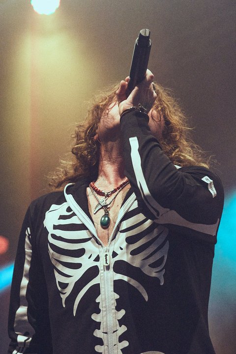 Ein Sänger mit langen, lockigen Haaren trägt einen schwarzen Hoodie mit einem Skelettdesign. Er hält ein Mikrofon und scheint begeistert zu singen. Die Szenerie ist von bunten Lichtern beleuchtet, die eine lebhafte Atmosphäre schaffen.