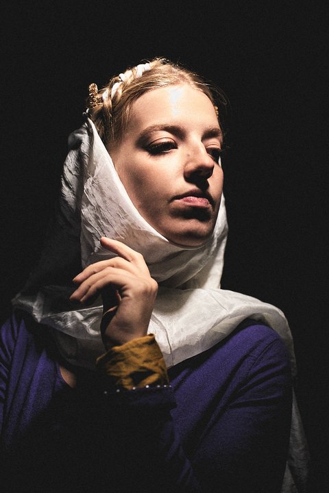 Eine Person mit einem hellen Kopftuch und einem lila Oberteil steht im Fokus. Ihr Gesicht zeigt einen nachdenklichen Ausdruck, während sie sanft ihr Kopftuch berührt. Der dunkle Hintergrund verstärkt die Konturen und den Ausdruck der Person.