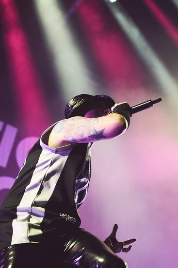 Ein Sänger steht auf der Bühne und hält ein Mikrofon. Er trägt ein schwarzes und weißes Outfit und hat sichtbare Tattoos auf dem linken Arm. Bunte Bühnenlichter scheinen auf ihn, während er in einer dynamischen Pose performt.