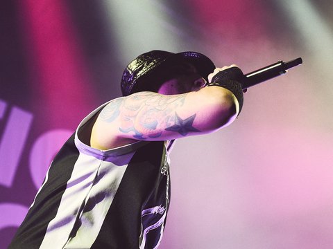 Ein Sänger steht auf der Bühne und hält ein Mikrofon. Er trägt ein schwarzes und weißes Outfit und hat sichtbare Tattoos auf dem linken Arm. Bunte Bühnenlichter scheinen auf ihn, während er in einer dynamischen Pose performt.