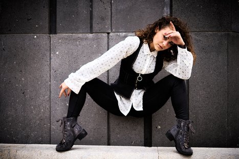 A person with curly hair poses in a crouched stance against a textured wall. They wear a black vest over a white, patterned shirt and black pants, completing the look with stylish boots. The pose conveys a sense of attitude and confidence.