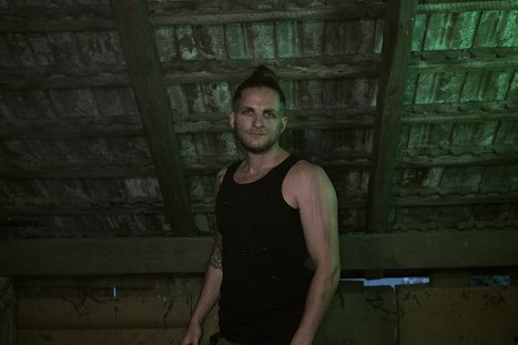 Ein Mann mit kurzen Haaren und einem schwarzen Tanktop steht unter einem rustikalen, schrägen Dach. Sein Blick ist ernst und direkt in die Kamera gerichtet. Die Beleuchtung hat einen grünen Farbton, der eine mysteriöse Atmosphäre schafft.