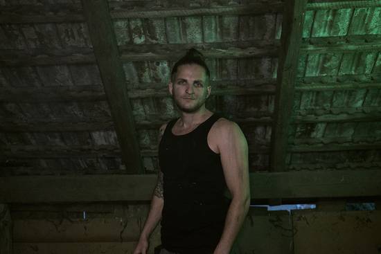 Ein Mann mit kurzen Haaren und einem schwarzen Tanktop steht unter einem rustikalen, schrägen Dach. Sein Blick ist ernst und direkt in die Kamera gerichtet. Die Beleuchtung hat einen grünen Farbton, der eine mysteriöse Atmosphäre schafft.