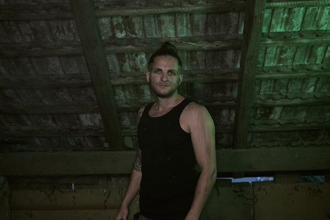 Ein Mann mit kurzen Haaren und einem schwarzen Tanktop steht unter einem rustikalen, schrägen Dach. Sein Blick ist ernst und direkt in die Kamera gerichtet. Die Beleuchtung hat einen grünen Farbton, der eine mysteriöse Atmosphäre schafft.