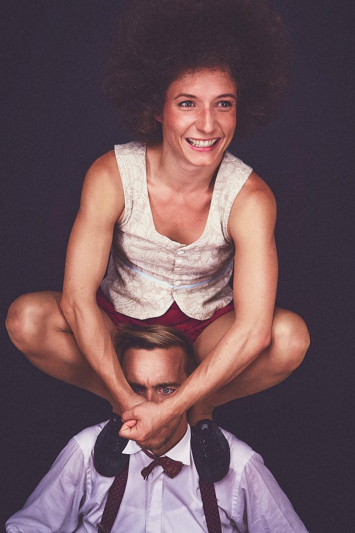 Eine Frau mit lockigem Haar sitzt auf den Schultern eines Mannes und grinst. Sie trägt ein ärmelloses Oberteil und Shorts, während der Mann einen Anzug und Fliege trägt. Beide scheinen eine fröhliche und humorvolle Atmosphäre zu schaffen.