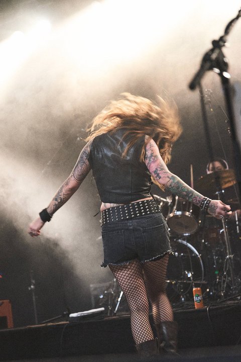 Eine rockende Musikerin mit langen, welligen Haaren und farbenfrohen Tattoos dreht sich auf der Bühne. Sie trägt eine schwarze Lederjacke und einen kurzen Jeansrock mit Netzstrümpfen. Im Hintergrund sind Schlagzeug und andere Bandmitglieder zu erkennen, während Scheinwerferlicht die Szene beleuchtet.